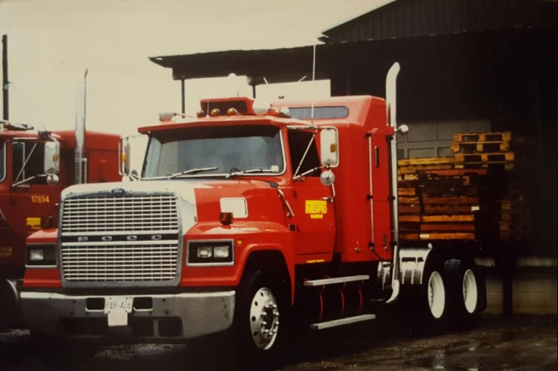 A large red Ford semi-truck with chrome details is parked near a building stacked with wooden pallets. Another similar red truck is partially visible in the background. The scene appears overcast and wet.