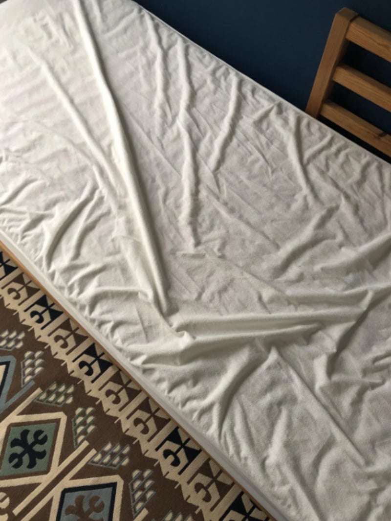 A white bedsheet with noticeable wrinkles forming a large letter "V" lies on a mattress next to a wooden chair and a patterned rug.