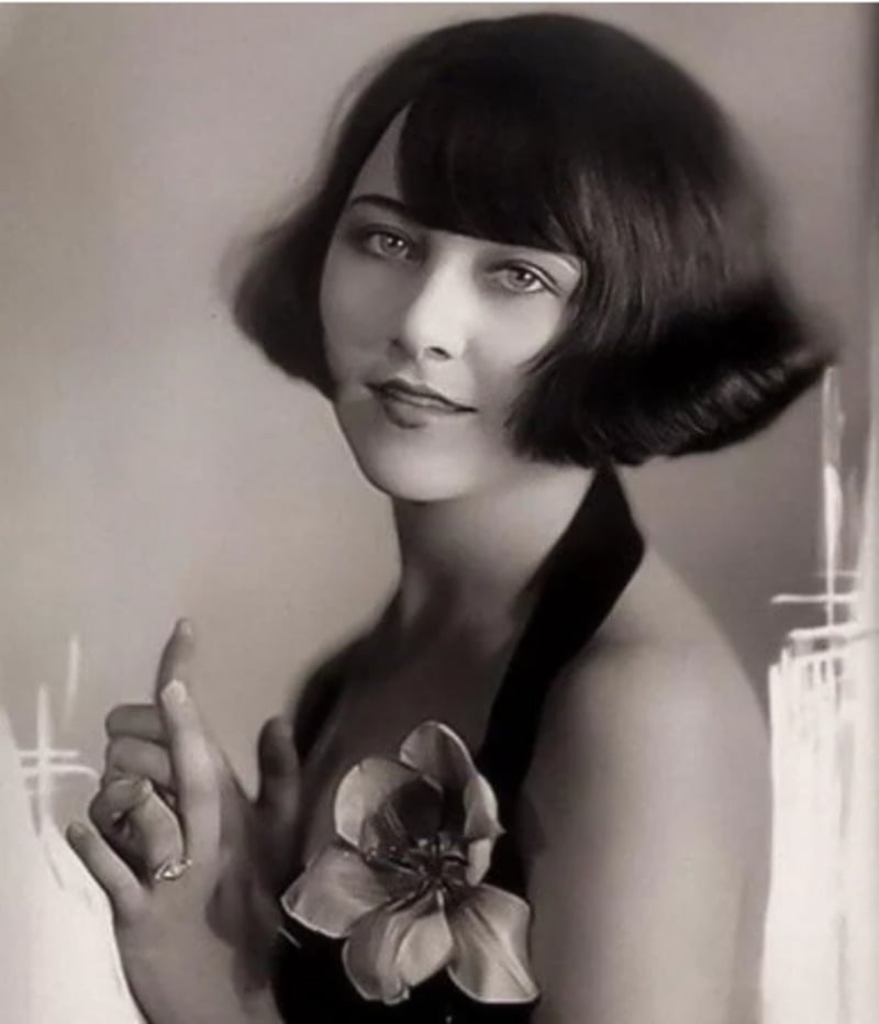 A woman with short, dark bobbed hair wears a halter dress adorned with a large flower and looks softly at the camera, hands gently clasped, in a black-and-white vintage portrait.