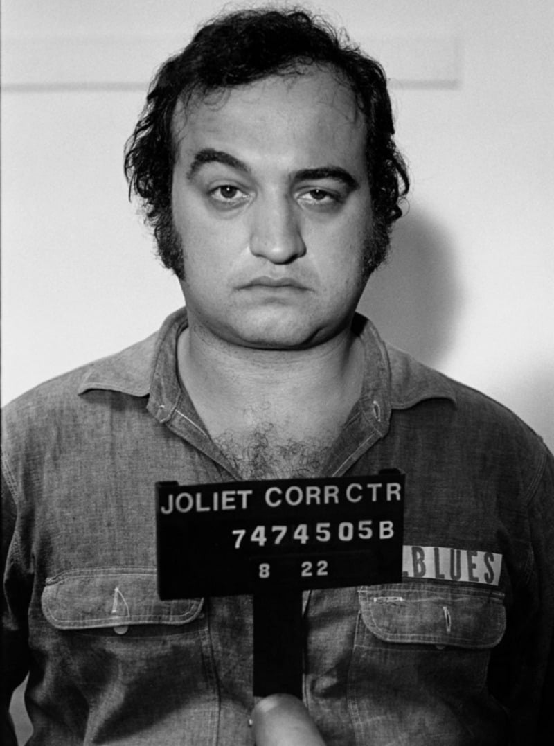 Black-and-white mugshot of a man with curly hair wearing a prison uniform labeled "BLUES," holding a sign that reads "Joliet Corr Ctr 7474505B." He has a neutral expression.