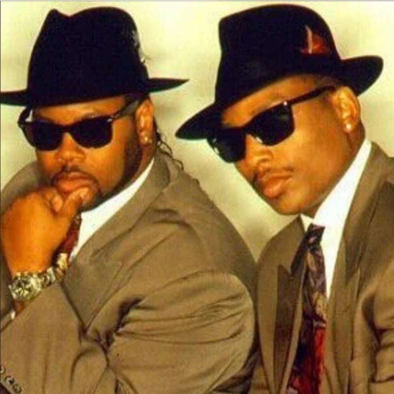 Two men wearing suits, ties, black hats, and dark sunglasses pose confidently against a plain background. One man rests his hand on his chin, while the other angles his head slightly, both with serious expressions.