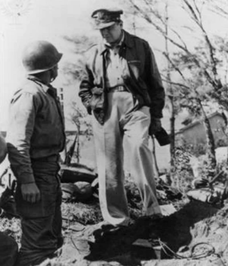 A military officer wearing a cap and jacket stands with one foot raised on a mound, looking down at a soldier in uniform and helmet. Trees and equipment are visible in the background.