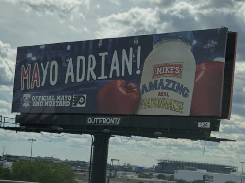 A billboard features a jar of “Mike’s Amazing Real Mayonnaise” with red boxing gloves and the words “Mayo Adrian!” mimicking “Yo Adrian!” The billboard is for the official mayo and mustard of the Philadelphia Flyers.