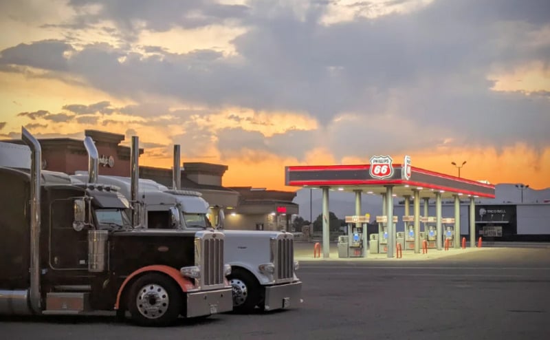Two semi trucks are parked in front of a Phillips 66 gas station at sunset, with an orange and blue sky in the background and the station lights brightly illuminating the fuel pumps.