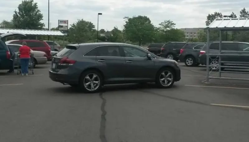 A dark gray car is parked diagonally across two parking spaces in a parking lot. Several other vehicles are parked nearby, and a person is loading groceries into a car. Trees and buildings are visible in the background.