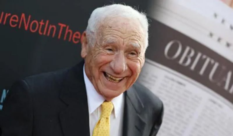 An elderly man in a suit and yellow tie smiles broadly. Behind him is a blurred newspaper with the word "OBITUARY" visible in large letters.