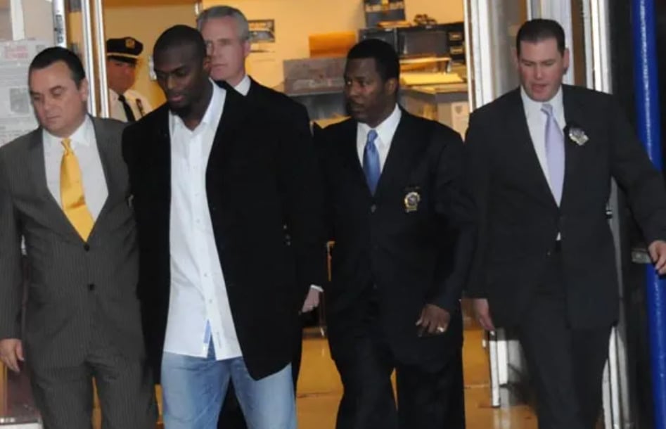 A group of five men, including one in handcuffs, are walking together indoors. Several men are wearing suits, and one appears to be a law enforcement officer escorting the man in handcuffs.