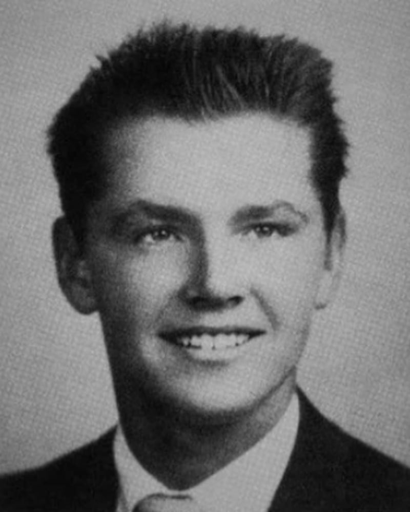 Black and white yearbook-style portrait of a young man with short, dark hair, smiling and wearing a suit jacket, dress shirt, and tie against a plain background.