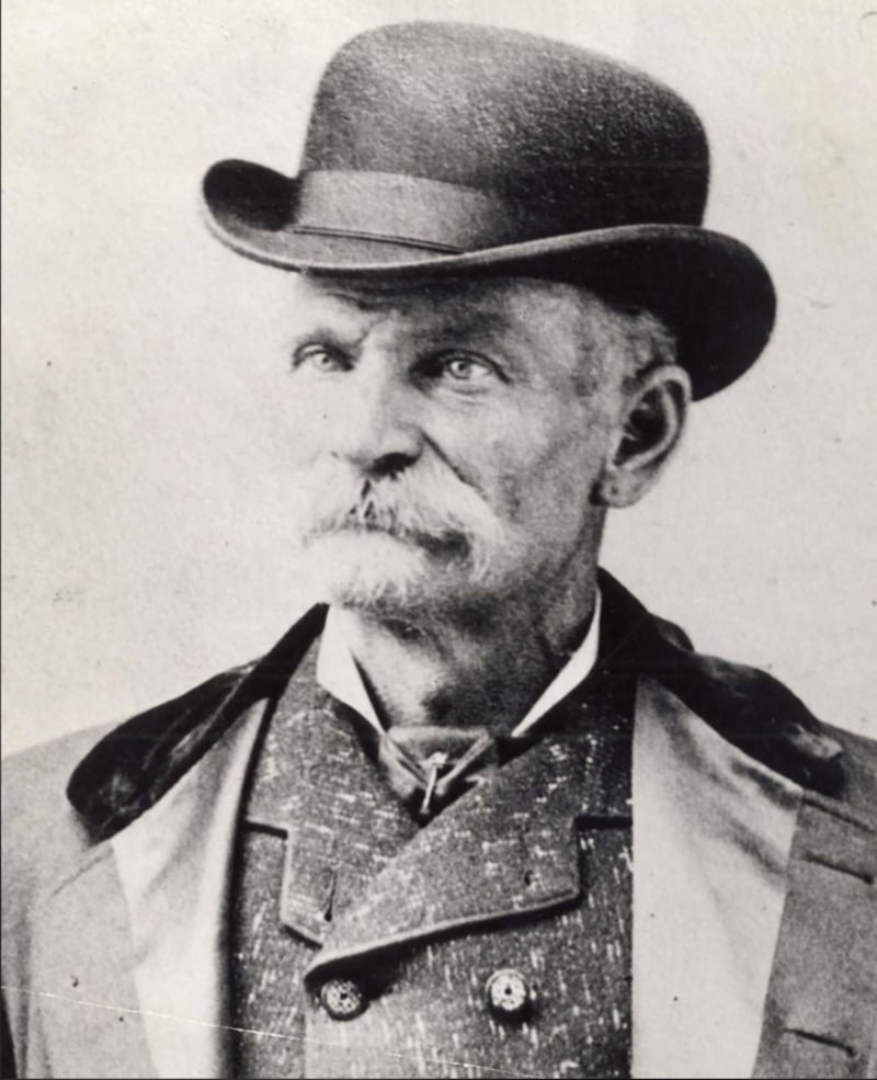 Black and white portrait of an older man with a mustache, wearing a bowler hat, a coat with wide lapels, and a high-collared shirt. He is looking slightly to the side with a serious expression.