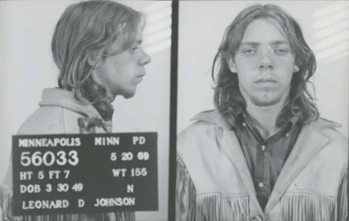 Black-and-white mugshot of a young man with long hair wearing a fringed jacket, shown in front and side profiles. A placard displays personal and booking information from Minneapolis Police, dated 1969.