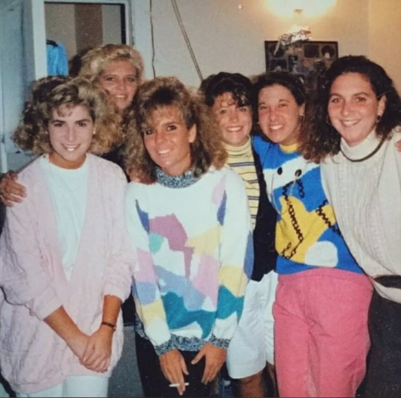 Six women stand closely together indoors, smiling at the camera. They wear colorful, patterned 1980s or 1990s-style sweaters, and a few have voluminous, curly hair typical of that era. The atmosphere seems cheerful and friendly.