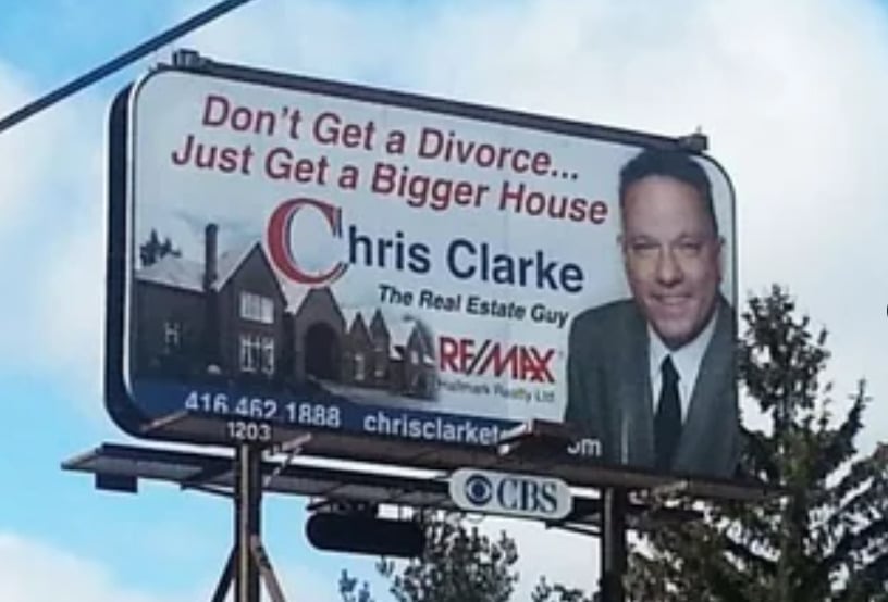 A billboard shows a smiling man in a suit with the text: "Don’t Get a Divorce... Just Get a Bigger House. Chris Clarke, The Real Estate Guy, RE/MAX." There is a photo of a large house and contact info below.