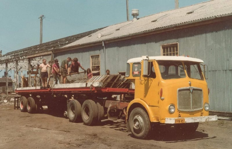 A yellow flatbed truck is parked beside a corrugated metal building. Several people stand on the truck’s trailer, loading or unloading metal sheets and other materials. The setting appears industrial or construction-related.