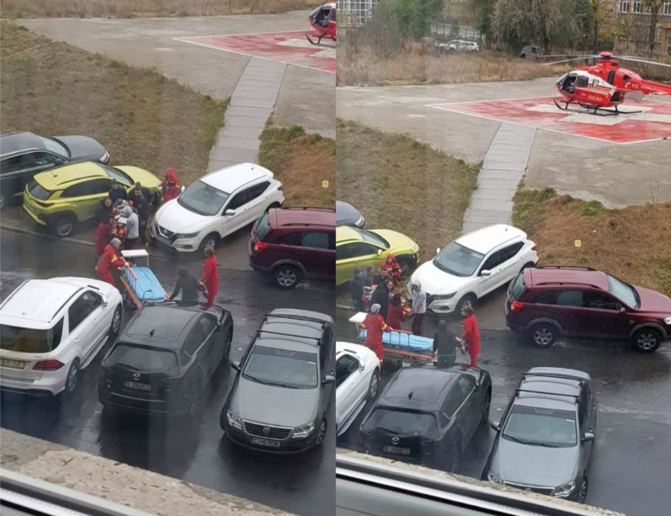 Paramedics in red uniforms carry a patient on a stretcher toward a rescue helicopter on a helipad. Several parked cars and emergency personnel are visible on the street beside the helipad.