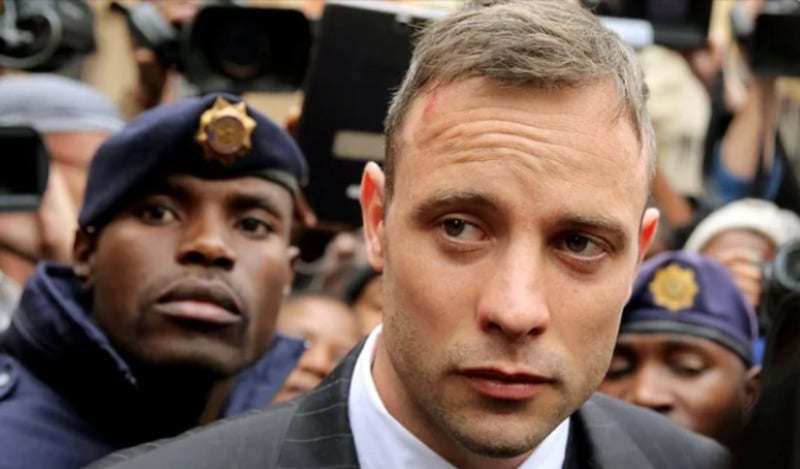 A man in a suit with a serious expression stands among police officers and reporters, with cameras and microphones visible in the background. One officer in uniform and beret is close beside him.
