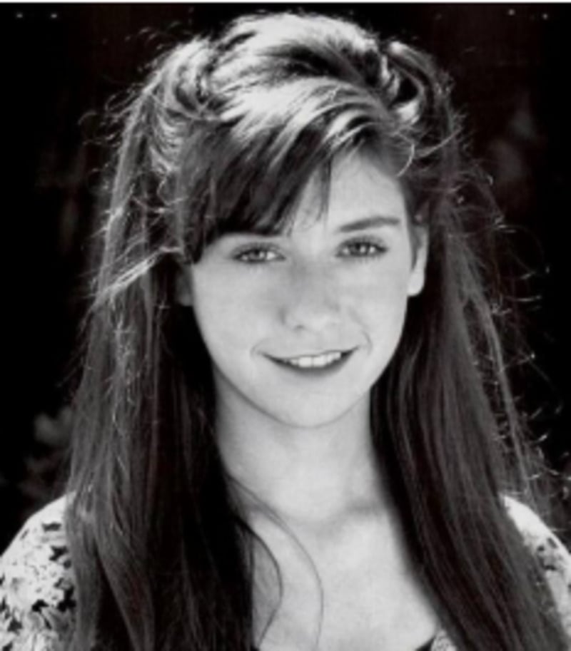 Black and white portrait of a young woman with long, dark hair and bangs, smiling softly at the camera. She is wearing a patterned top and is positioned against a blurred, dark background.