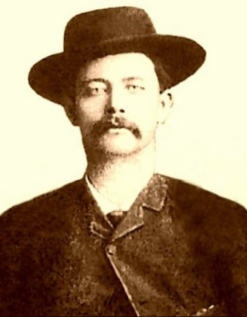 Sepia-toned portrait of a man with a mustache wearing a wide-brimmed hat, a dark suit jacket, and a tie, looking directly at the camera against a plain background.