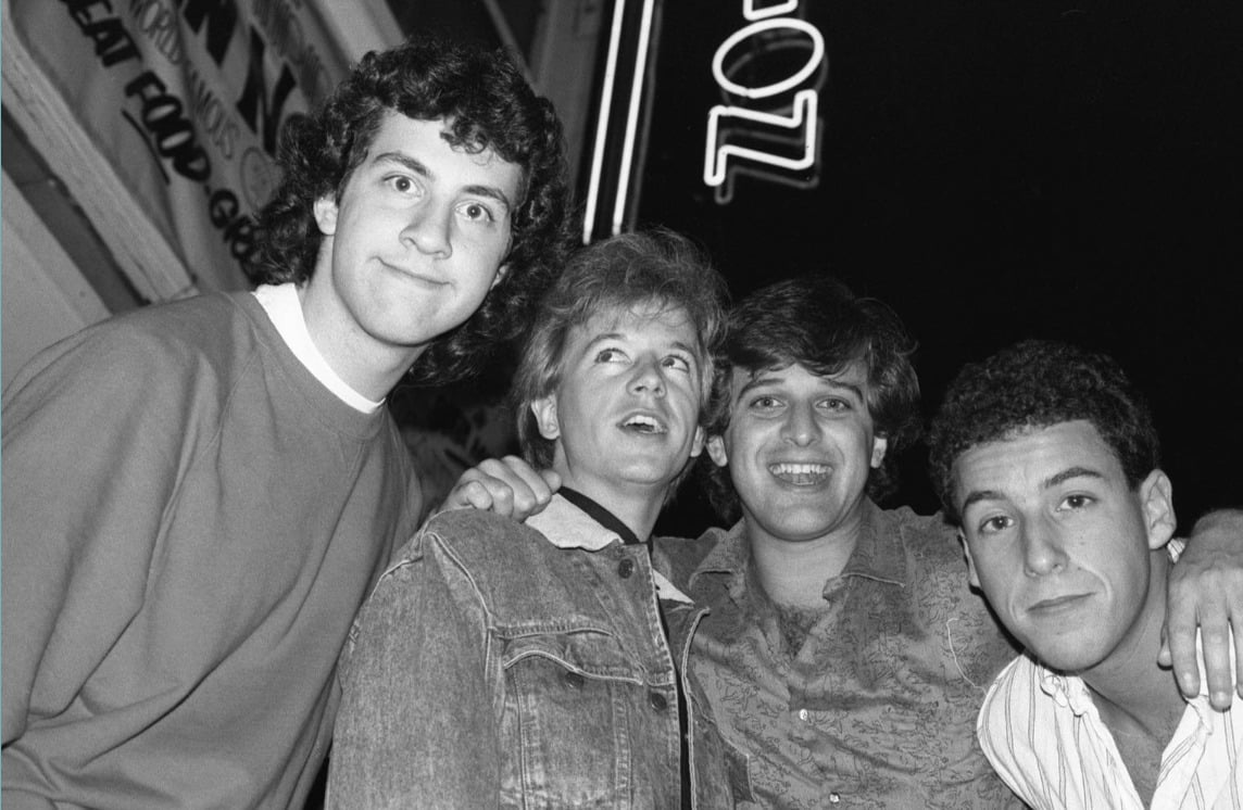 Four young men pose together and smile for a black-and-white photo at night, standing close under a neon sign. One has his arm around another, and they all appear to be enjoying themselves.