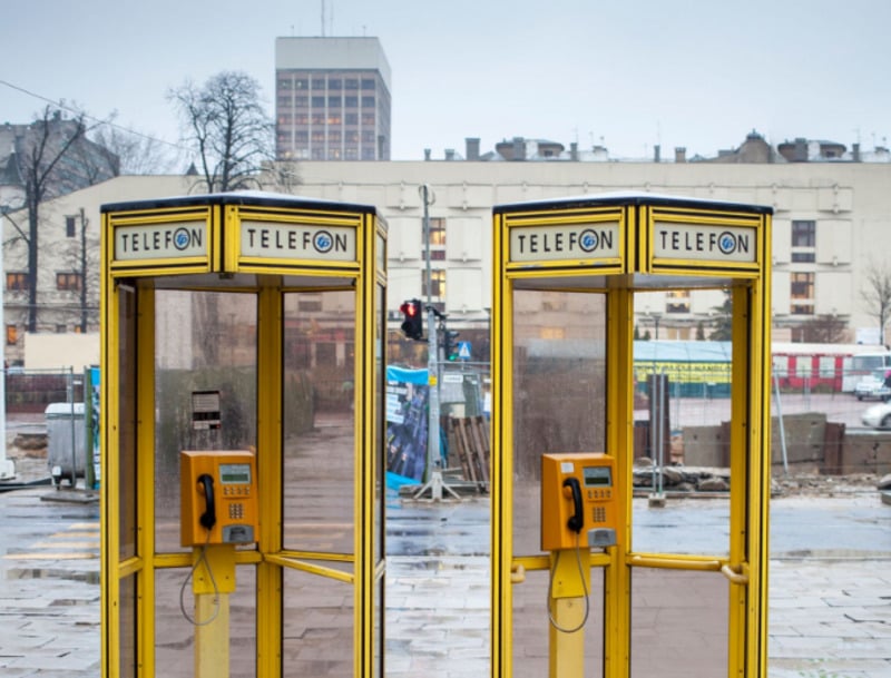Two yellow phone booths with the word "TELEFON" on top stand side by side on a paved sidewalk in a city, with buildings, construction, and a cloudy sky in the background.