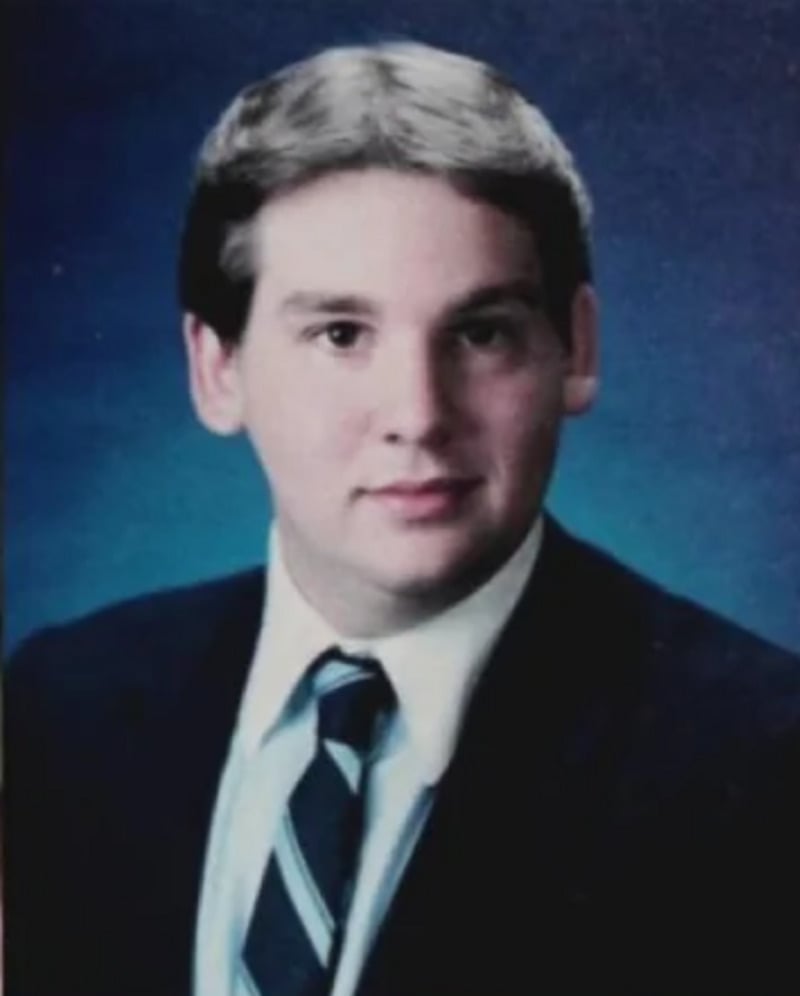 A young man with short, light brown hair wears a dark suit, white shirt, and striped tie, posing for a formal portrait against a blue background.