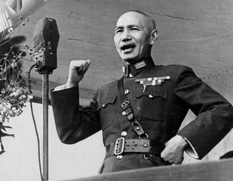 A man in a military uniform speaks passionately into a vintage microphone, raising his right fist while standing at a podium.
