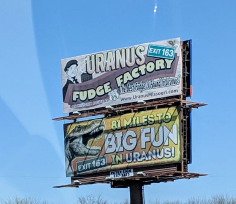 A roadside billboard advertises the Uranus Fudge Factory and reads “81 Miles to Big Fun in Uranus!” with a cartoon dinosaur and route directions, promoting a tourist stop in Missouri.