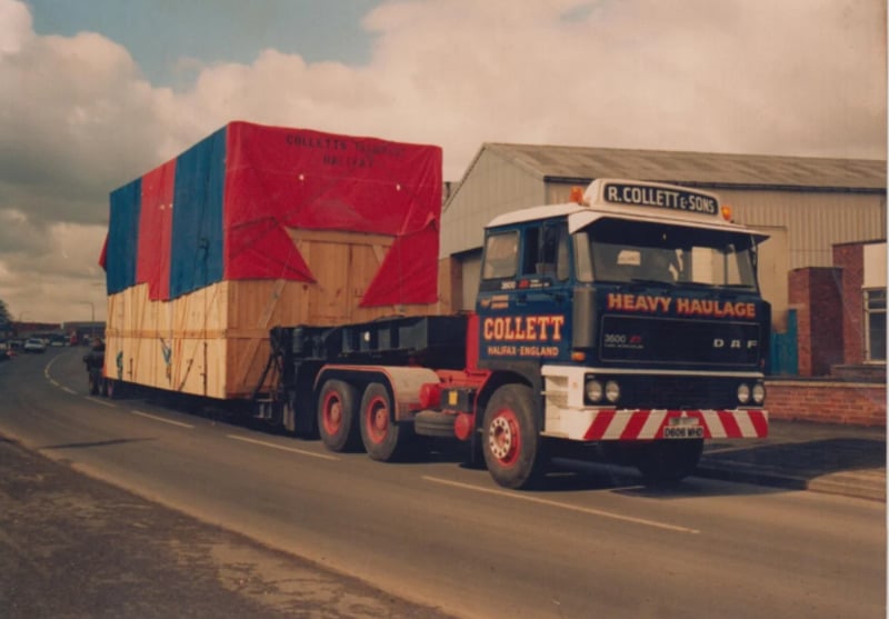 A large truck labeled “Heavy Haulage” transports a massive, tarped wooden crate on a road beside industrial buildings under a cloudy sky.