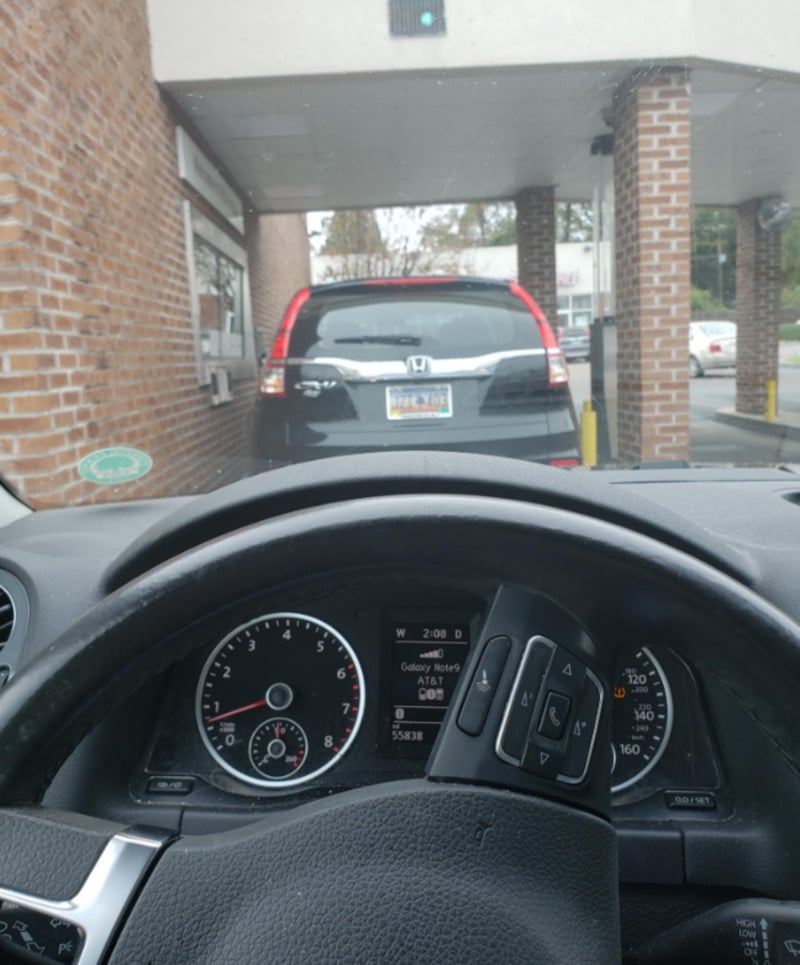 View from inside a car at a drive-thru, showing the dashboard, steering wheel, and a Honda SUV ahead in line by a brick building. The time on the car’s display reads 2:06 PM.