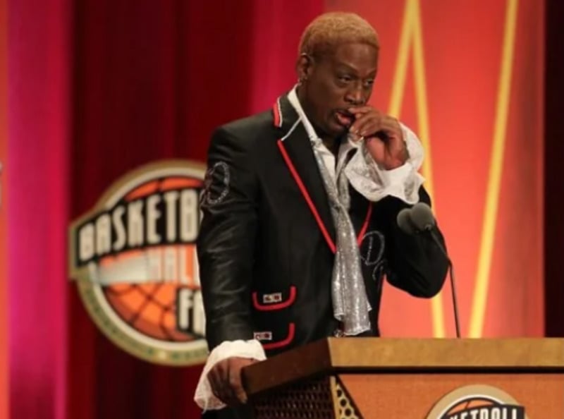 A man with blond hair, wearing a black suit with red trim and a glittery silver scarf, stands at a podium labeled "Basketball Hall of Fame," appearing emotional while speaking into a microphone.