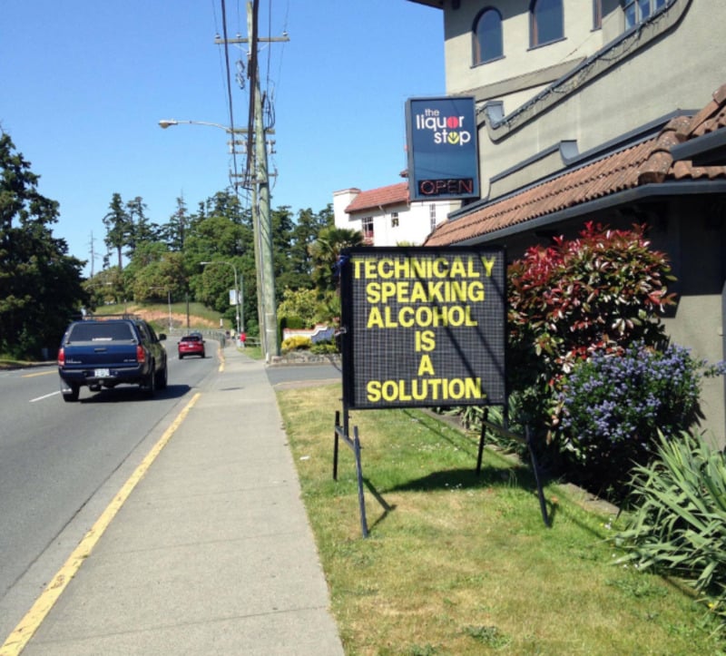 A sidewalk sign outside a liquor store reads, "Technically speaking, alcohol is a solution." Cars and greenery line the street beside the store.