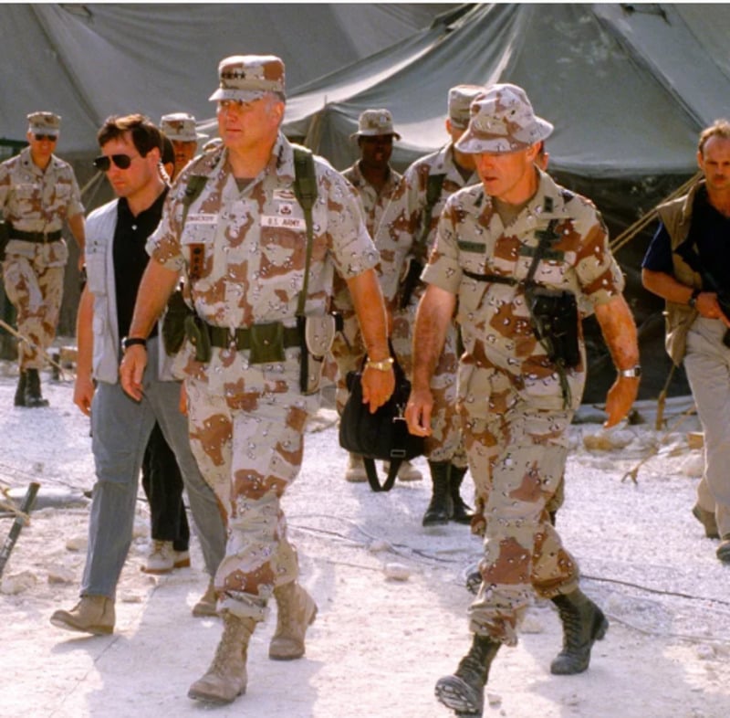 Group of military personnel in desert camouflage uniforms and gear walk outside near tents, accompanied by civilians. The environment appears dry and dusty, suggesting a desert or field camp location.