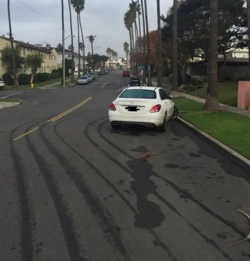 A white car is parked on a residential street with long, dark skid marks curving on the road behind it. Palm trees and houses line both sides of the street.