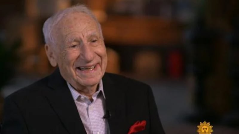An elderly man with short white hair smiles warmly, wearing a dark suit jacket, light button-up shirt, and red pocket square. The background is softly blurred, with a yellow sun logo at the bottom right corner.