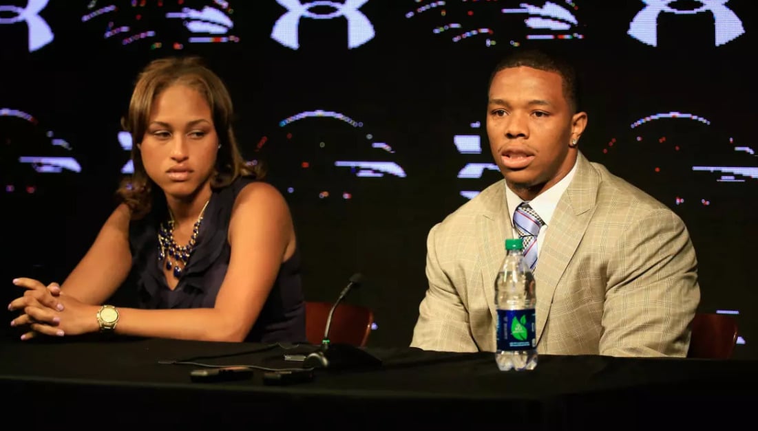 A woman and a man sit side by side at a table with microphones and a water bottle, speaking at a press conference with a black backdrop displaying the Under Armour logo.