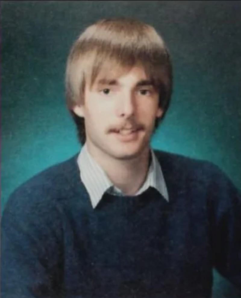 A young man with light brown hair and a mustache poses for a formal portrait. He is wearing a blue sweater over a collared shirt against a teal studio background.