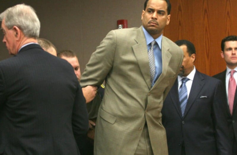 A man in a tan suit is being handcuffed by an officer in a courtroom, surrounded by several other men in suits. The mood appears serious and tense.