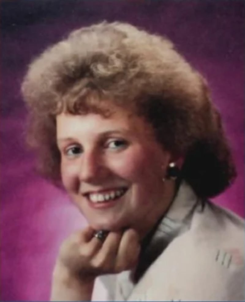 A woman with curly, light brown hair smiles at the camera against a purple background. She rests her chin on her hand and wears a light-colored top and small, dark earrings.