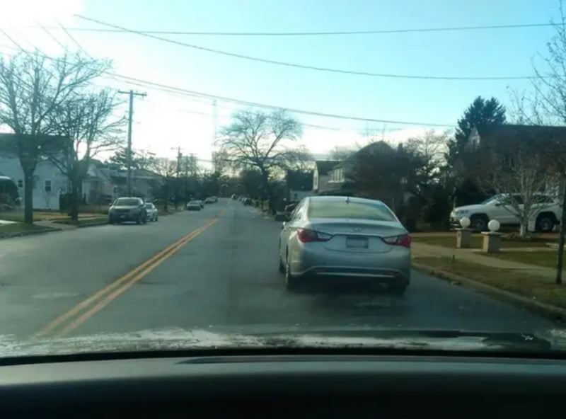 A suburban street view from inside a car, showing another silver car driving ahead. The road is lined with houses, leafless trees, and parked cars under a clear sky.