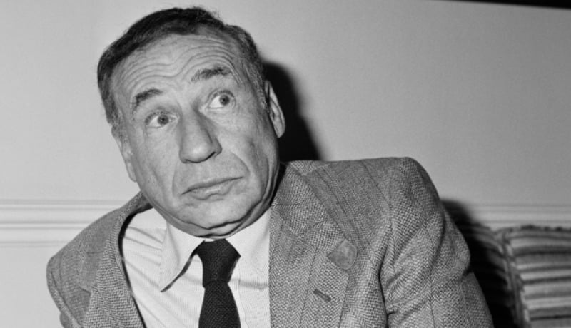 A man in a suit and tie sits, looking to the side with a surprised or puzzled expression. The image is black and white, and he is indoors against a plain background.