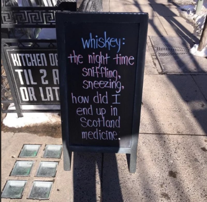 A sidewalk chalkboard sign reads: "whiskey: the night time sniffling, sneezing, how did I end up in Scotland medicine." Snow and sidewalk are visible around the sign.