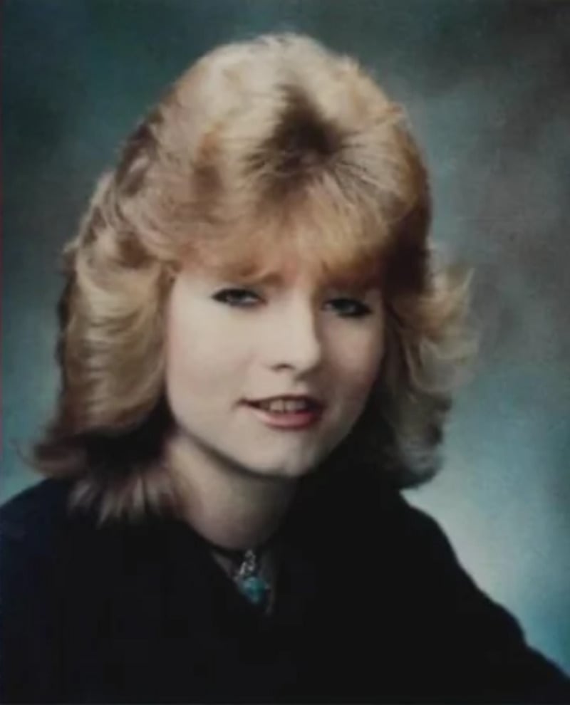 A young woman with feathered, shoulder-length blonde hair poses for a formal portrait against a blurred blue-gray background. She is wearing a black top and a necklace, and is smiling slightly.