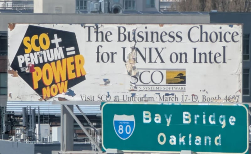 A weathered billboard advertises "SCO + Pentium = Power Now" and "The Business Choice for UNIX on Intel" above a green highway sign for Bay Bridge and Oakland, with buildings visible in the background.
