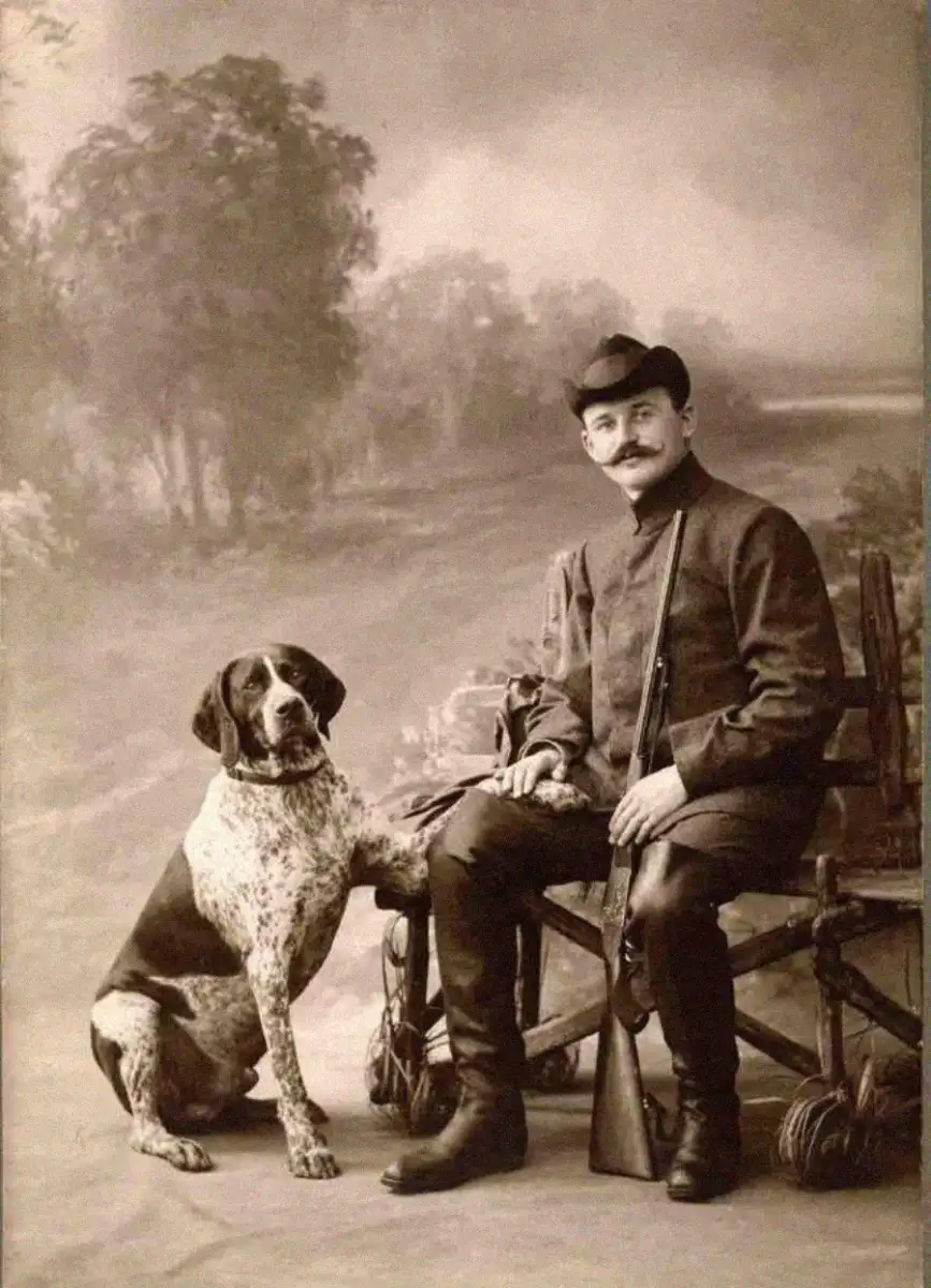 A vintage black-and-white photo of a man in hunting attire sitting on a wooden bench, holding a rifle, with a large dog sitting beside him, and a painted outdoor backdrop behind them.