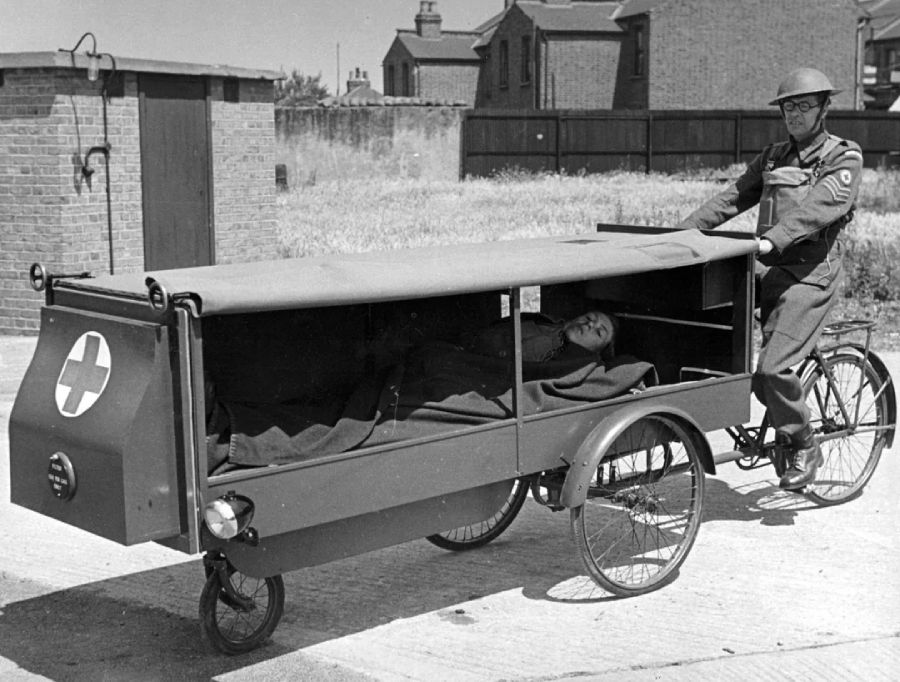 A soldier in uniform stands next to a three-wheeled bicycle ambulance with a covered compartment, transporting a patient lying inside, on a paved area near brick buildings.