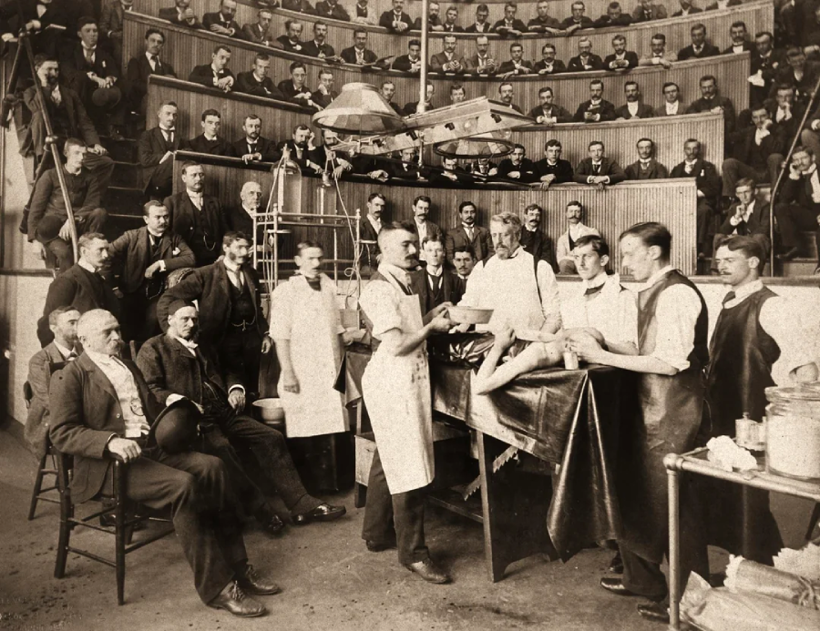 A black-and-white photo shows surgeons performing an operation on a patient in an old-fashioned operating theater, as numerous men in suits observe from tiered seats.