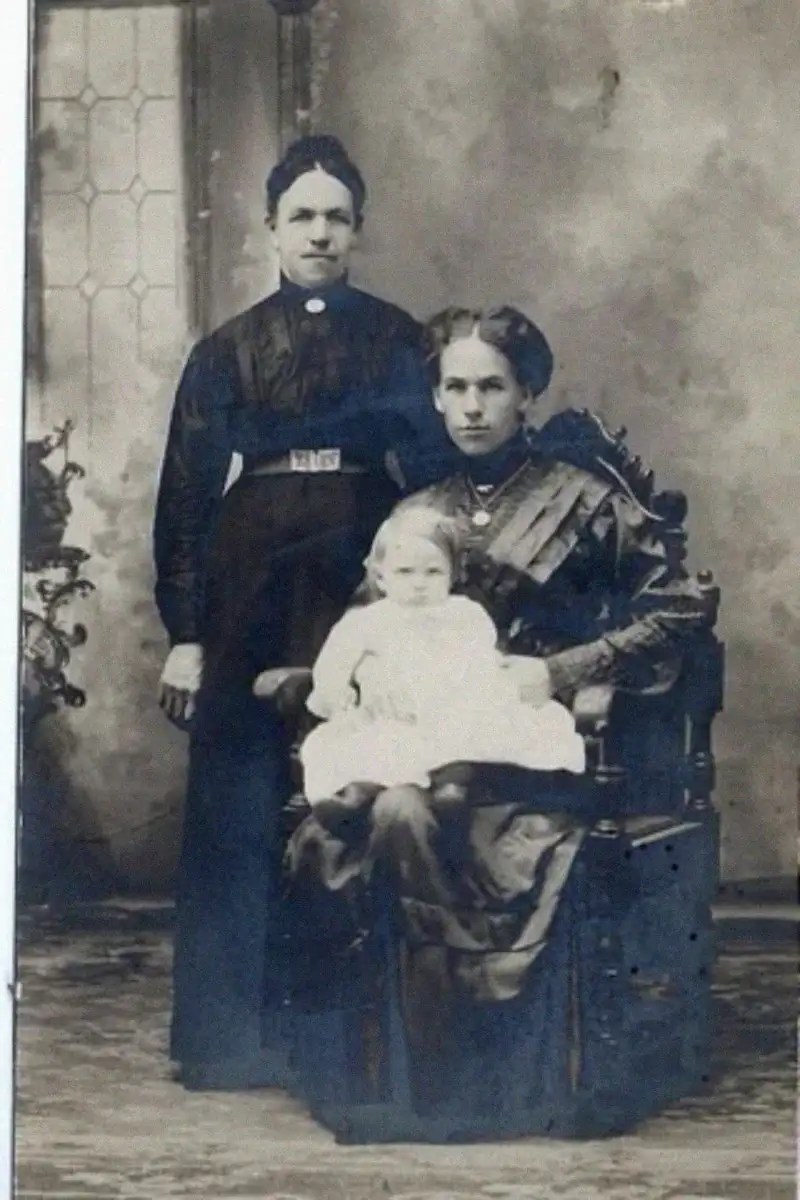 Three people pose for a vintage black-and-white portrait: a woman stands behind another seated woman holding a small child in a white dress, all wearing formal clothing from the early 1900s.
