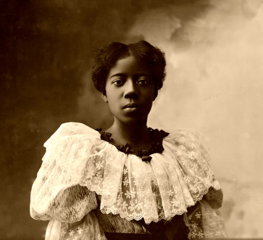 A young Black woman with natural hair styled up, wearing a formal dress with large puffed lace sleeves, poses for a sepia-toned portrait against a softly blurred studio backdrop.