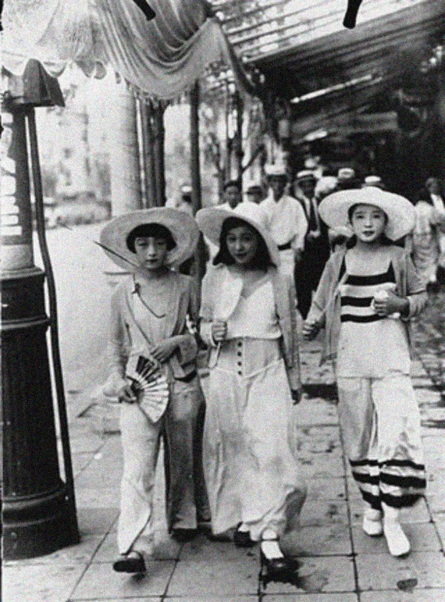 Three young women walk side by side down a city sidewalk, dressed in vintage clothing and wide-brimmed hats, with one holding a fan. Other pedestrians are visible in the background.