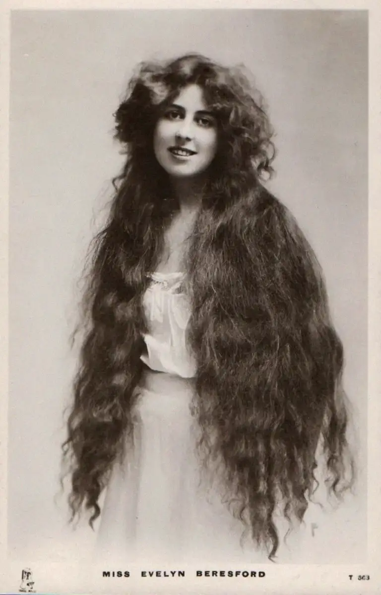 A woman with very long, wavy hair wears a light blouse and skirt, smiling at the camera. The text at the bottom reads "Miss Evelyn Beresford." The photo is in black and white.