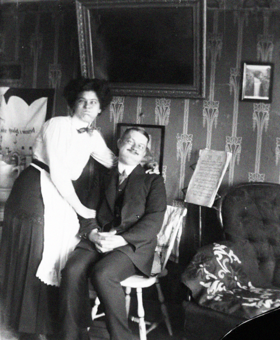 A vintage black-and-white photo of a woman standing beside a seated man in a decorated room with patterned wallpaper, a chair, framed pictures, and a sheet of music in the background.